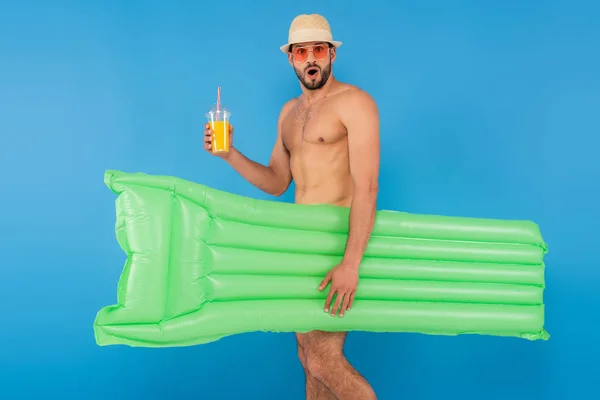 Hombre sin camisa excitado sosteniendo el jugo de naranja y el colchón inflable aislados en azul - foto de stock