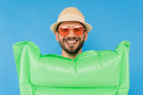 Улыбающийся мужчина в солнечной шляпе и солнцезащитных очках возле надувного матраса, изолированного на голубом — стоковое фото