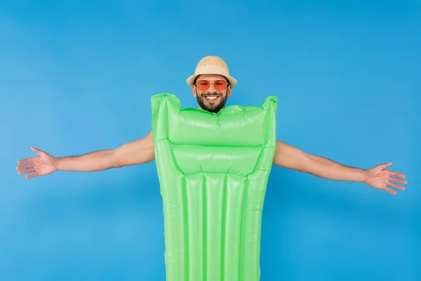 Hombre sonriente en sombrero de sol y gafas de sol de pie cerca de colchón inflable aislado en azul — Stock Photo