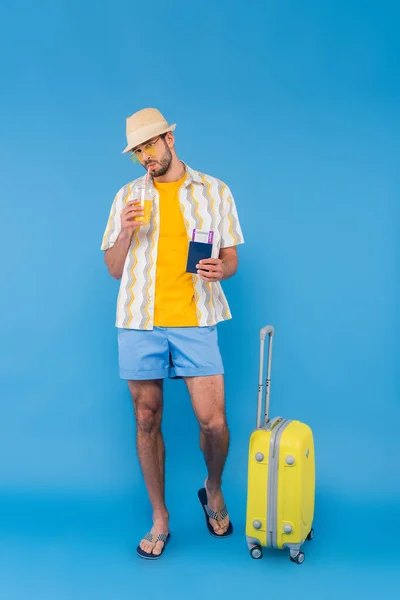 Man in sunglasses drinking orange juice and holding passport near suitcase on blue background — Stock Photo