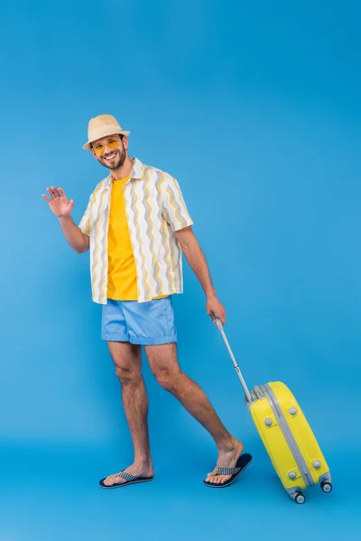 Smiling man in sunglasses waving hand and holding suitcase on blue background — Stock Photo