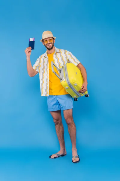 Cheerful man in sun hat holding passport and suitcase on blue background — Stock Photo