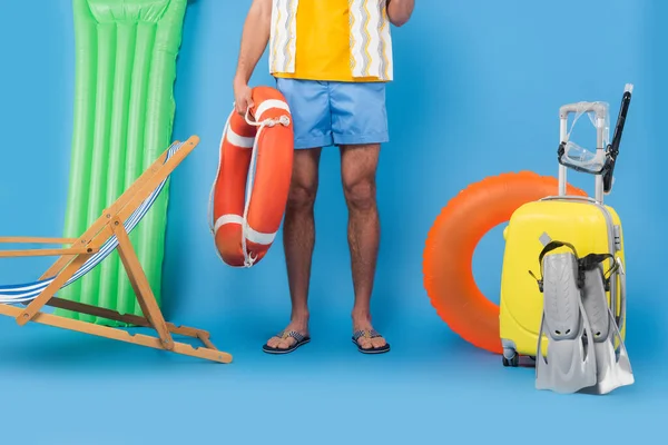 Vista recortada del hombre sosteniendo boya de vida cerca de la silla de cubierta, maleta y aletas de natación sobre fondo azul - foto de stock