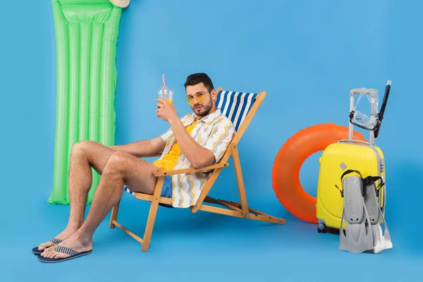 Man in sunglasses holding orange juice on deck chair near inflatable ring and suitcase on blue background — Stock Photo