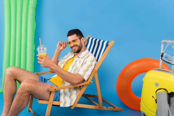 Hombre sonriente con jugo de naranja sentado en la silla de cubierta cerca de la maleta, aletas de natación y anillo inflable sobre fondo azul - foto de stock