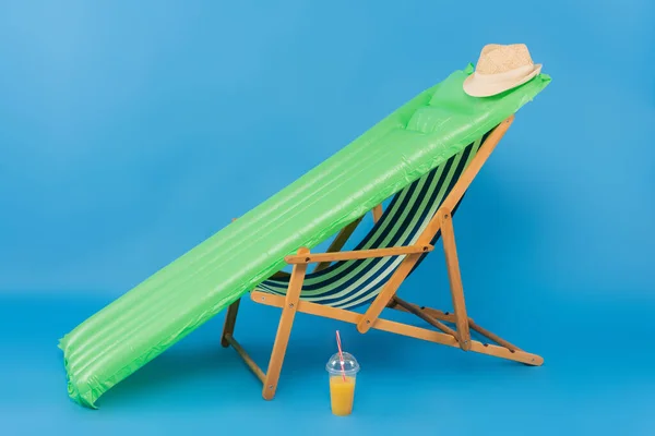 Sombrero solar en colchón inflable cerca de la silla de cubierta y jugo de naranja sobre fondo azul - foto de stock