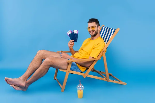 Smiling man holding passports while sitting on deck chair near orange juice on blue background — Stock Photo
