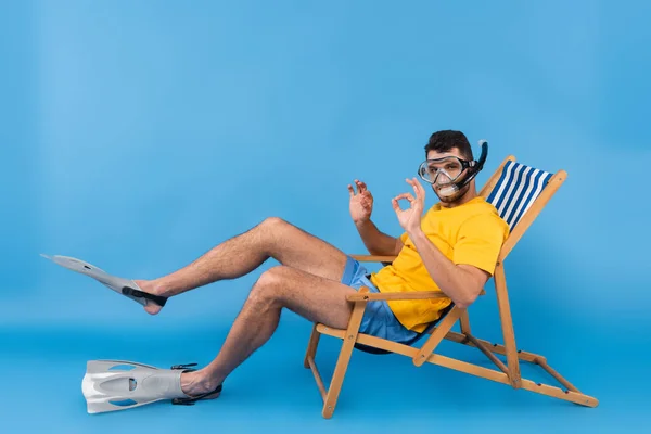 Homem em óculos de natação e nadadeiras mostrando gesto ok na cadeira de praia em fundo azul — Fotografia de Stock