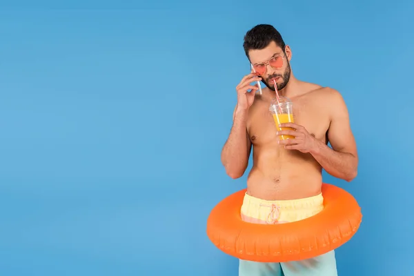 Shirtless man in sunglasses and inflatable ring drinking orange juice and talking on smartphone isolated on blue — Stock Photo