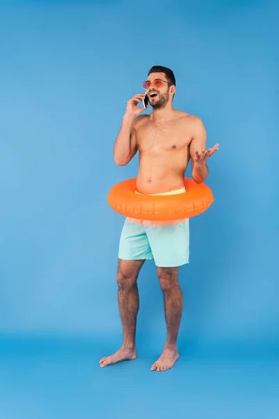 Hombre alegre sin camisa en anillo inflable y gafas de sol hablando en el teléfono inteligente sobre fondo azul - foto de stock