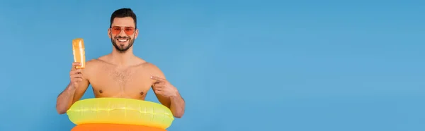 Homme souriant avec anneaux gonflables pointant vers la crème solaire isolée sur bleu, bannière — Photo de stock