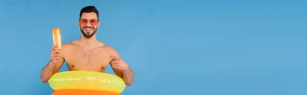 Smiling man with inflatable rings pointing at sunscreen isolated on blue, banner — Stock Photo