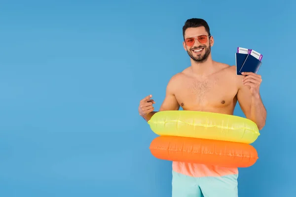 Hombre positivo en gafas de sol y anillos inflables apuntando a pasaportes aislados en azul - foto de stock