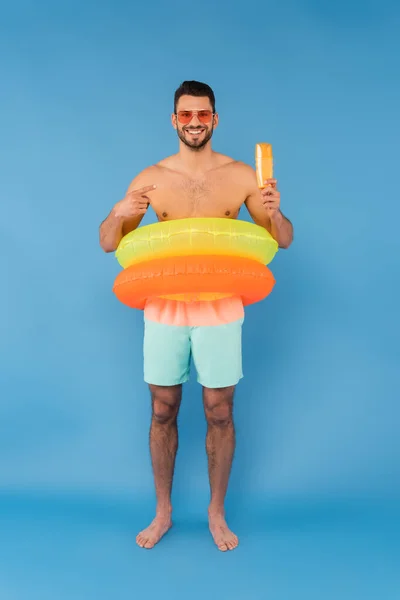 Hombre sin camisa en anillos inflables apuntando al protector solar sobre fondo azul - foto de stock