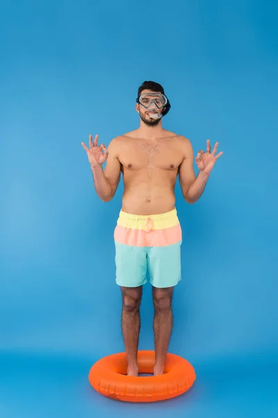 Hombre sin camisa en gafas de natación mostrando gesto aceptable mientras está de pie en el anillo inflable sobre fondo azul - foto de stock