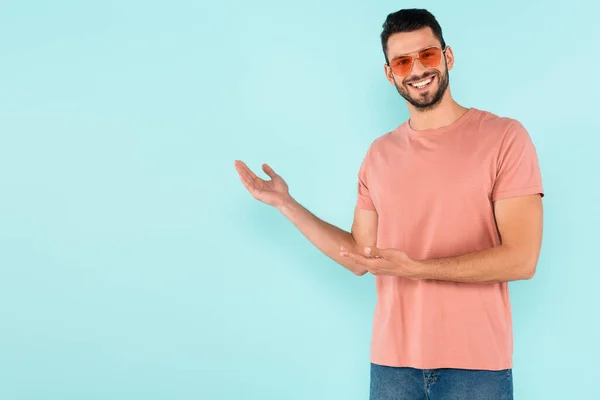 Lächelnder Mann mit Sonnenbrille zeigt mit den Händen auf blauem Grund — Stockfoto