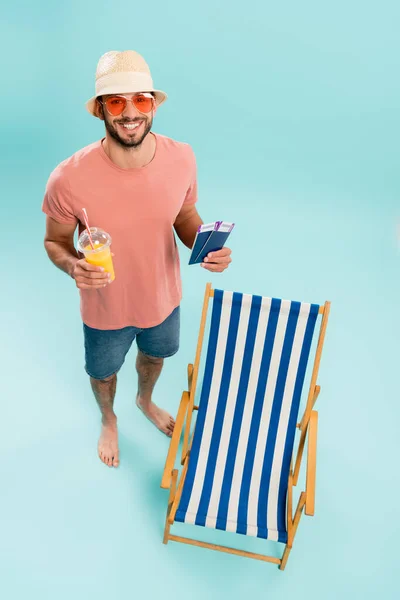 Vista de ángulo alto del hombre positivo en gafas de sol que contienen jugo de naranja y pasaportes cerca de la silla de cubierta sobre fondo azul - foto de stock