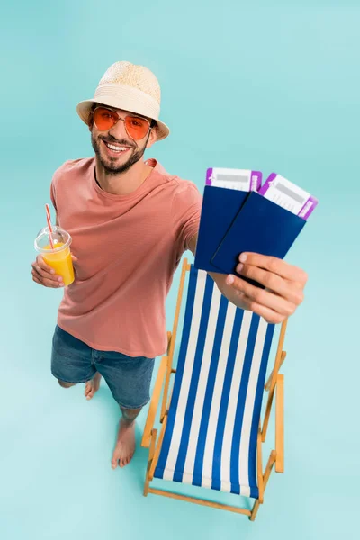 Vista de alto ángulo de pasaportes borrosos en manos del hombre sonriente con jugo de naranja cerca de la silla de cubierta sobre fondo azul - foto de stock