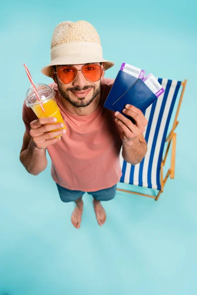 Vista superior del hombre en gafas de sol que contienen jugo de naranja y pasaportes con billetes de avión cerca de la silla cubierta borrosa sobre fondo azul - foto de stock