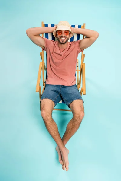 Vista de ángulo alto del hombre positivo en sombrero de sol y gafas de sol sentado en la silla de cubierta sobre fondo azul - foto de stock