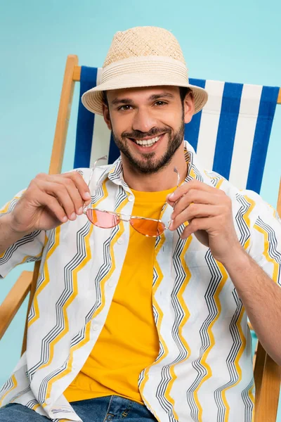 Hombre positivo en sombrero de paja sosteniendo gafas de sol en la silla de cubierta aislado en azul - foto de stock
