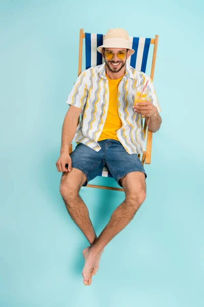 Vista de ángulo alto del hombre feliz en sombrero de paja que sostiene el jugo de naranja en la silla de cubierta sobre fondo azul - foto de stock