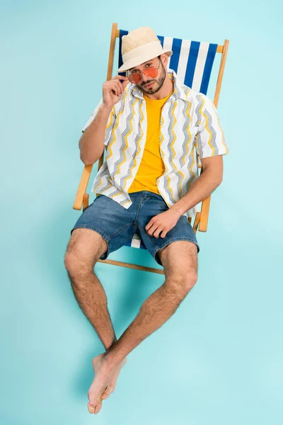 Vista de alto ángulo del hombre barbudo en gafas de sol y sombrero de paja mirando a la cámara sobre fondo azul - foto de stock