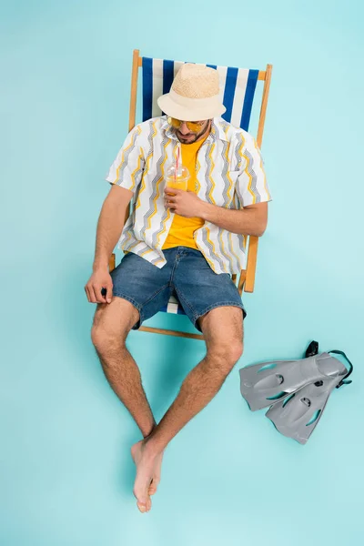 High angle view of man in straw hat holding orange juice near swimming flippers on blue background — Stock Photo