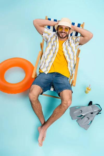 High angle view of happy man in sunglasses sitting on deck chair near inflatable ring and swimming flippers on blue background — Stock Photo