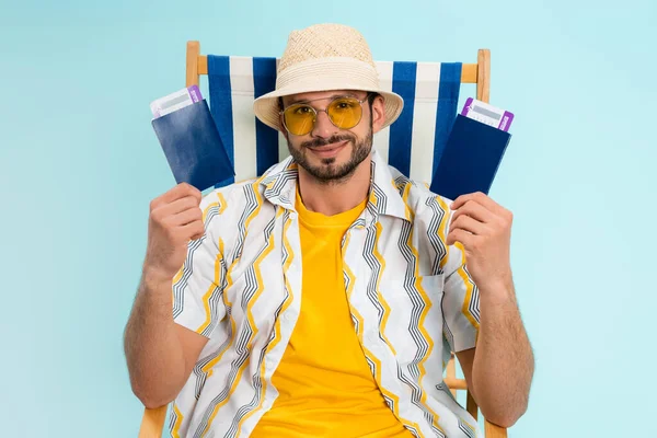 Hombre sonriente con sombrero de paja sosteniendo pasaportes con boletos de avión en la silla de cubierta aislado en azul - foto de stock