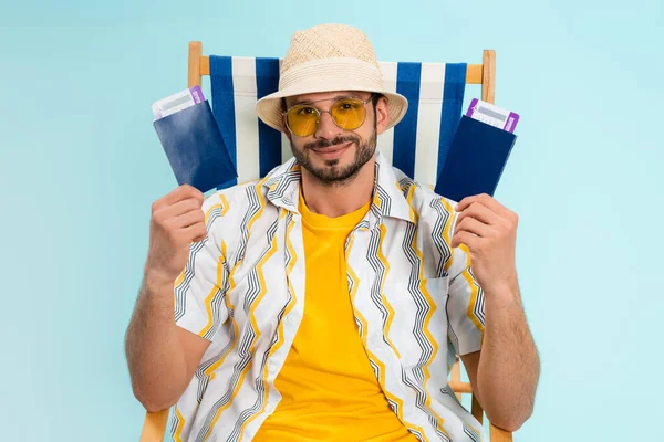 Lächelnder Mann mit Strohhut und Pässen mit Flugtickets auf Liegestuhl isoliert auf blauem Grund — Stockfoto