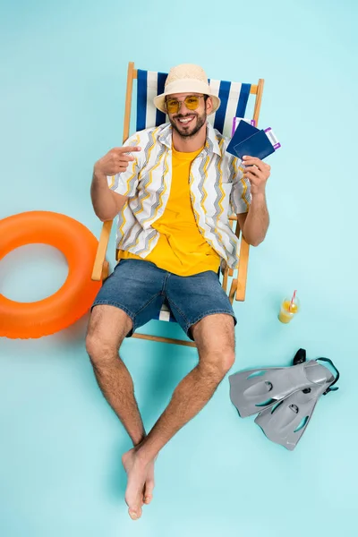 Vue grand angle de l'homme souriant pointant vers les passeports près des nageoires, du jus d'orange et de l'anneau gonflable sur fond bleu — Photo de stock