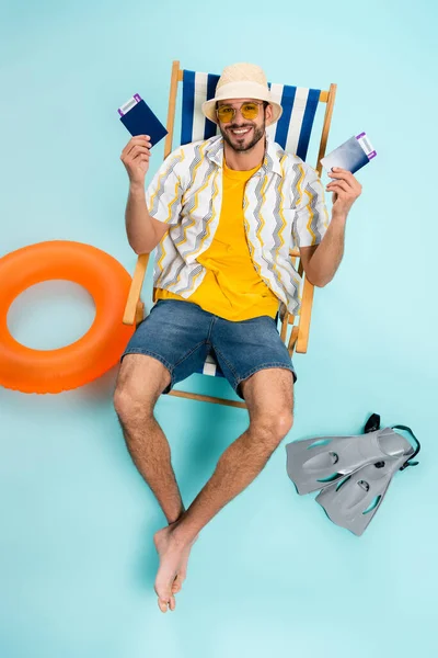 Vue grand angle de l'homme souriant dans des lunettes de soleil tenant passeports près des nageoires et anneau gonflable sur fond bleu — Photo de stock