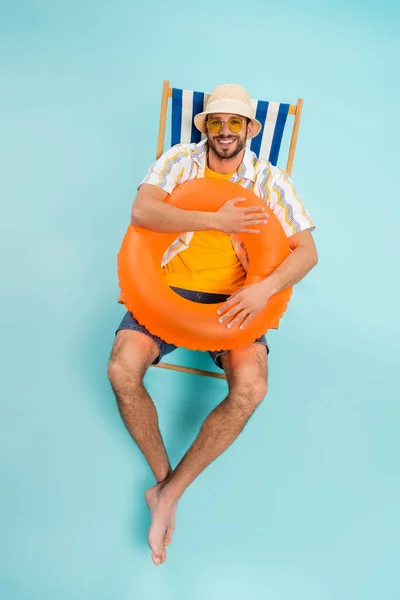 High angle view of positive man in straw hat holding inflatable ring on deck chair on blue background — Stock Photo