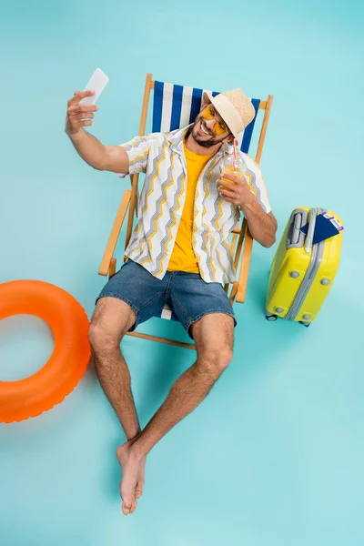 Vista de ángulo alto del hombre sonriente con jugo de naranja tomando selfie cerca del anillo inflable y la maleta con pasaportes sobre fondo azul - foto de stock
