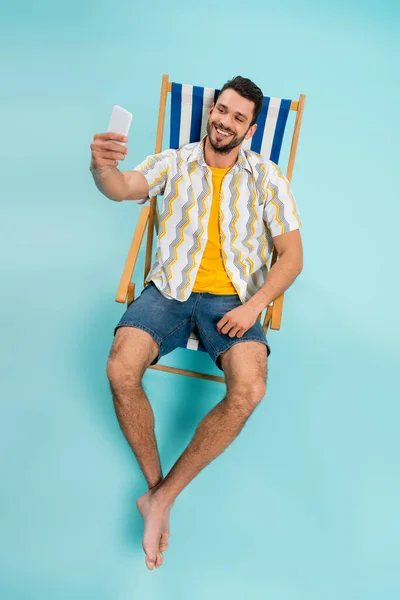 Vista de ángulo alto del hombre sonriente tomando selfie mientras está sentado en la silla de cubierta sobre fondo azul - foto de stock