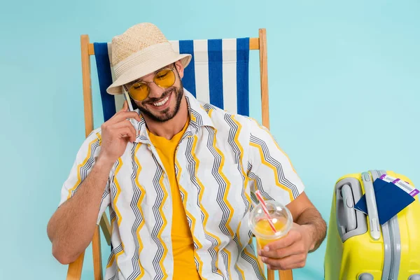 Positive man talking on smartphone and holding orange juice near suitcase and passports on blue background — Stock Photo