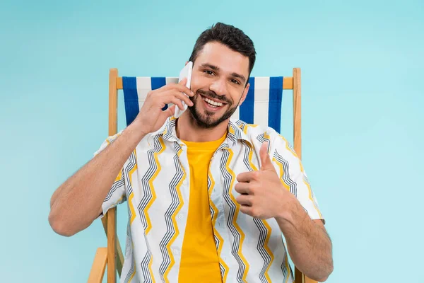Sonriente hombre mostrando como mientras habla en el teléfono inteligente en la silla de cubierta aislado en azul - foto de stock