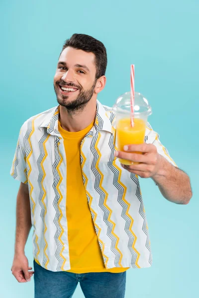 Sorrindo homem segurando suco de laranja desfocado em copo de plástico isolado em azul — Fotografia de Stock