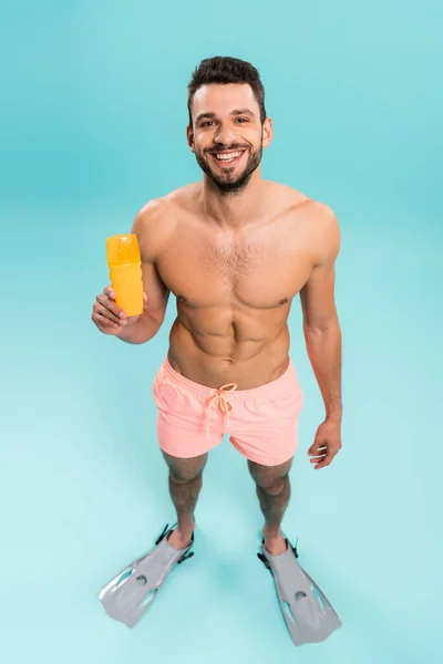 Vista de ángulo alto del hombre sin camisa en aletas de natación sosteniendo protector solar sobre fondo azul - foto de stock