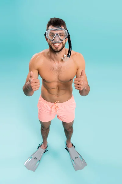 High angle view of shirtless man in swimming goggles and flippers showing like on blue background — Stock Photo