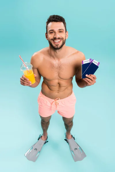 Vista de ángulo alto del hombre sin camisa positivo sosteniendo jugo de naranja y pasaportes sobre fondo azul - foto de stock