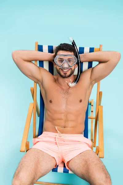 Positive shirtless man in swimming goggles sitting on deck chair on blue background — Stock Photo