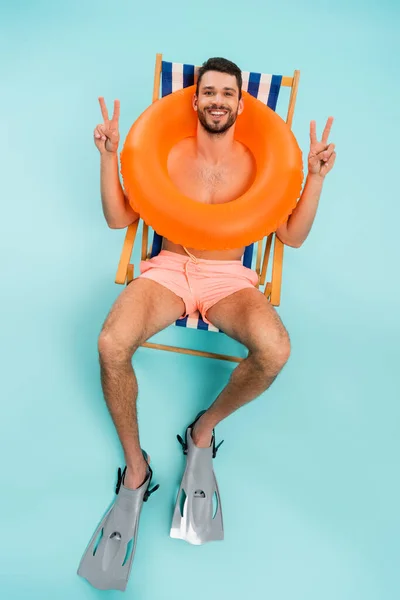 Vue grand angle de l'homme torse nu dans l'anneau gonflable et nage palmes montrant la paix sur fond bleu — Photo de stock