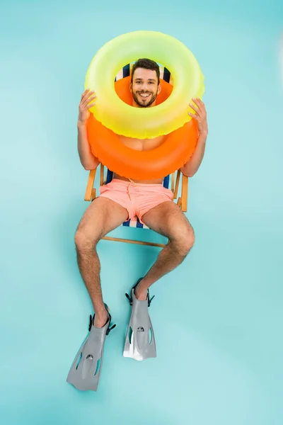 Vue grand angle de l'homme souriant dans les nageoires et les anneaux gonflables assis sur la chaise longue sur fond bleu — Photo de stock