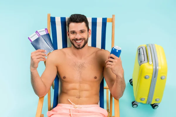 Uomo sorridente senza maglietta con bevande in scatola e passaporti vicino alla valigia su sfondo blu — Foto stock