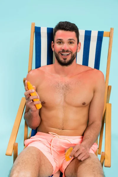 Positive man with sunburn holding sunscreen on deck chair isolated on blue — Stock Photo
