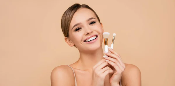 Mujer alegre sonriendo mientras posando con cepillos cosméticos aislados en beige, pancarta - foto de stock