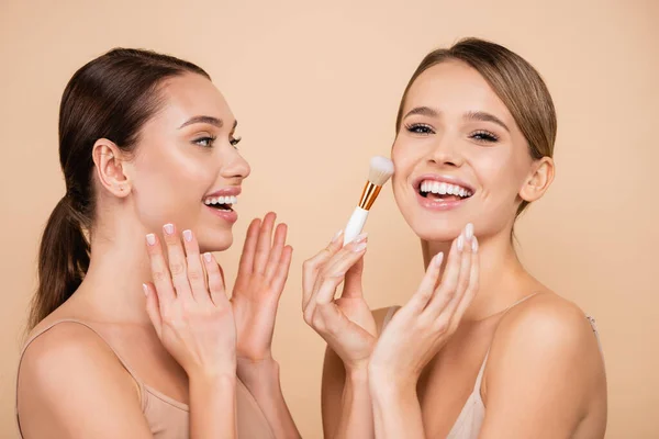 Amazed woman looking at laughing friend powdering face isolated on beige — Stock Photo