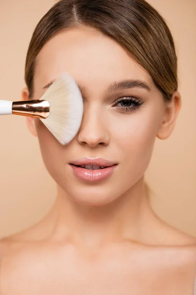 Pretty woman with natural visage covering eye with cosmetic brush isolated on beige — Stock Photo